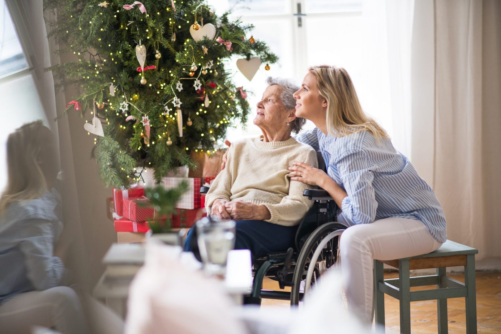 An elderly woman in a wheelchair and a blonde woman sitting next to each other. Both are looking at a Christmas tree and smiling.