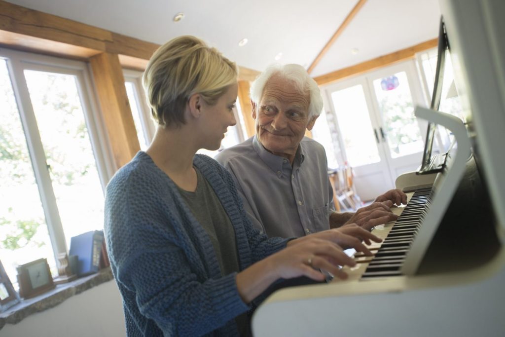 An elderly man and his carer playing the piano together. They are smiling at each other.